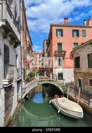 Small canal with an arched stone bridge and a covered boat between historic buildings in Venice Stock Photo