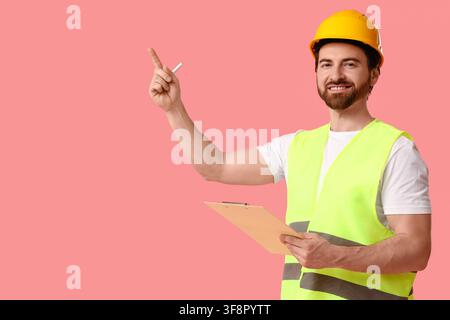 Young happy male construction worker in uniform with clipboard pointing at something on pink background Stock Photo