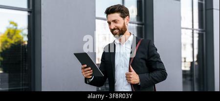 Smiling Man In Business Suit Using Tablet Outside Modern Office Building, Business Concept Stock Photo