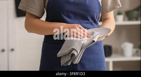 Person in a blue apron drying a clean white plate with a gray linen towel in a kitchen setting, Essence of domestic chores and kitchen hygiene Stock Photo