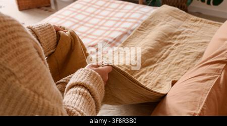 Person in a cozy sweater holding a soft, textured blanket on a bed with plaid bedding, Comfort and relaxation Stock Photo