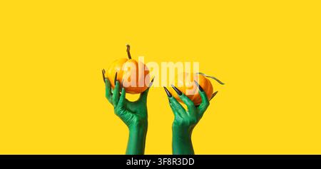 Green monster hands with long black nails holding two small orange pumpkins on a bright yellow background for halloween, this halloween themed image f Stock Photo