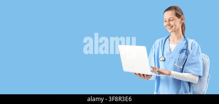 Smiling young female medical student in scrubs with a stethoscope and backpack using a laptop for online learning Stock Photo