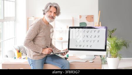 Smiling senior man with gray hair and beard using a graphic tablet for calligraphy design on a computer monitor, He is engaged in a creative hobby or Stock Photo