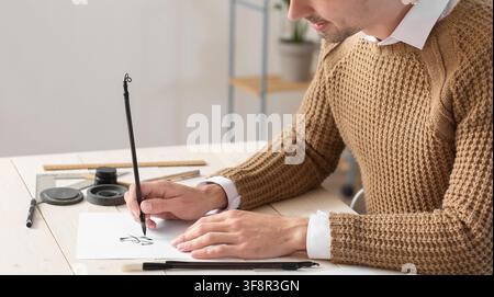 Young man practicing calligraphy with a traditional brush and ink on white paper at a wooden desk, a young man practices calligraphy, demonstrating ar Stock Photo