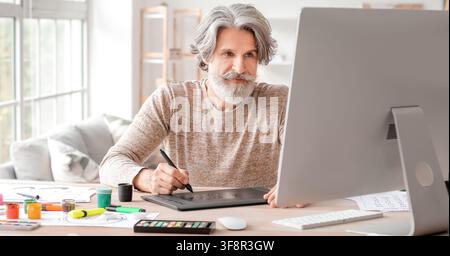 Mature man with gray hair and beard using a graphic tablet and computer for digital art and design work at home, He uses a graphic tablet and a comput Stock Photo