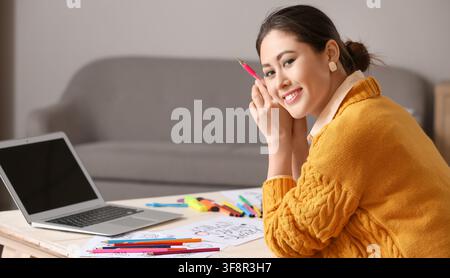 Smiling young asian woman holding a pink pen while working on creative projects at a desk with a laptop and art supplies Stock Photo