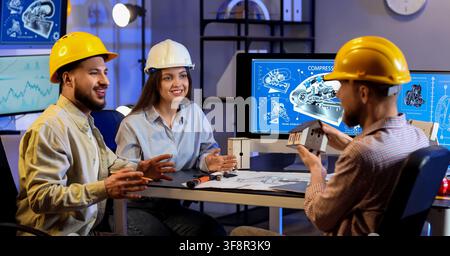 Three smiling engineers in hard hats discussing a house model and mechanical blueprints on computer screens in a modern office Stock Photo