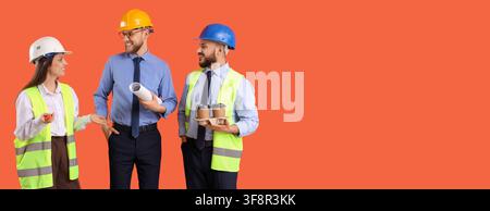 Three smiling construction engineers in hard hats and safety vests discussing plans with coffee on an orange background, One engineer holds rolled blu Stock Photo