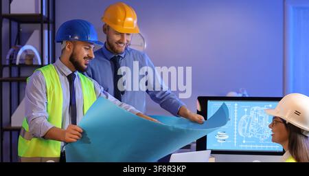 Smiling engineers in hard hats reviewing blueprints and technical drawings on a computer screen in an office, the team collaborates on a project, disc Stock Photo