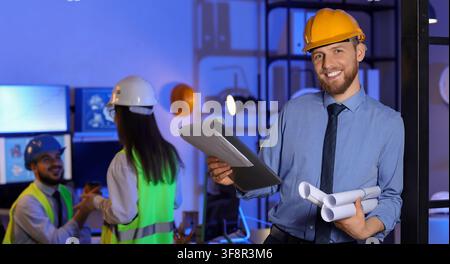 Smiling male engineer in a hard hat holding blueprints and a clipboard in a modern office with Colleagues, Colleagues in safety vests and hard hats ar Stock Photo