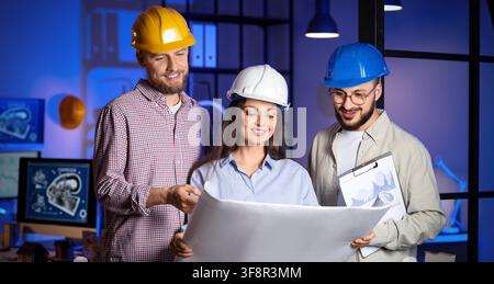 Three smiling engineers in hard hats reviewing blueprints and project plans in a modern office at night, the team is engaged in a collaborative discus Stock Photo