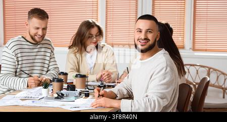 Smiling young man with beard looking at camera during a creative calligraphy and design workshop, He is seated at a table with other students, engaged Stock Photo