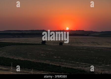Moon partially obscuring the sun during a solar eclipse over a vast rural landscape with agricultural fields, hills, and a vibrant orange sunset in Spain Stock Photo