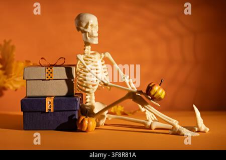 Seated skeleton holding mini pumpkin beside gift boxes on orange background for playful Halloween advertising or party poster Stock Photo