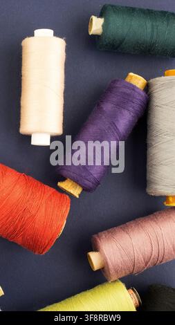 Variety of thread spools in different vibrant and pastel colors, including beige, orange, purple, green, and pink, spread across a textured dark blue Stock Photo