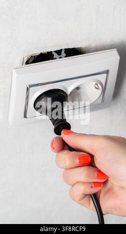 Close-up view of person hand with bright orange nail polish on fingers as plug electric cord into white wall socket. Socket partially dislodged, expos Stock Photo