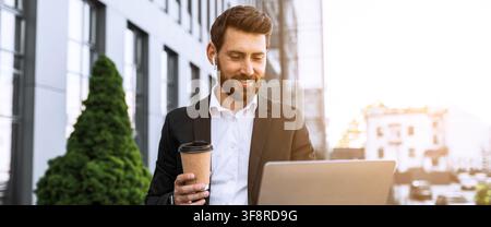 Happy Caucasian Man Using Laptop And Drinking Coffee Outdoors In Modern Business District Stock Photo