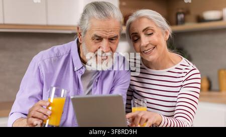 Happy Elderly Caucasian Couple Using Tablet Together In Kitchen, Enjoying Quality Time Stock Photo