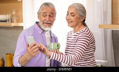 Happy Elderly Caucasian Couple Smiling Together In Kitchen, Using Smartphone And Drinking Coffee Stock Photo