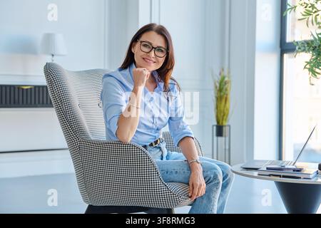 Smiling mature businesswoman in eyeglasses leaning on armchair in office Stock Photo