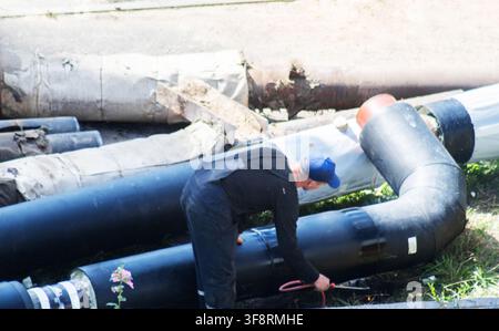 Adult worker in blue cap and dark uniform actively engaged in connection process of new heating pipeline system. Work involves large diameter black pi Stock Photo
