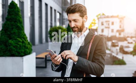 Smiling Caucasian Man Using Smartphone With Earphones In Urban Setting, Business Concept Stock Photo