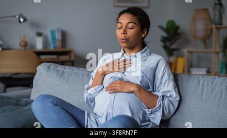 Pregnant Black Woman Practicing Breathing Exercises On Sofa At Home, Healthcare Concept Stock Photo