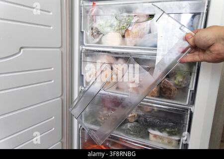 Close-up view of hand removing fractured transparent plastic storage tray from white freezer interior; contents of freezer, including various packaged Stock Photo