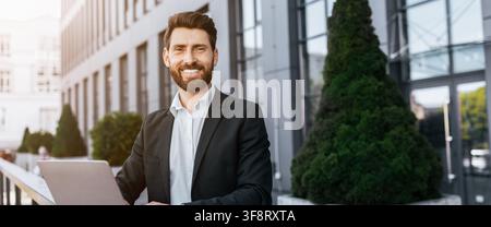 Smiling Young Caucasian Man Working On Laptop Outdoors In Business Setting Stock Photo