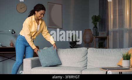 Happy Black Woman Arranging Decorative Pillows On Sofa In Modern Living Room, Home Interior Stock Photo