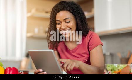 Happy Black Woman Using Tablet In Kitchen, Engaged And Smiling, Cooking Concept Stock Photo