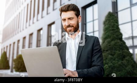 Smiling Caucasian Man Working On Laptop In Business Attire, Outdoor Office Setting Stock Photo