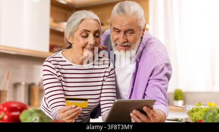 Happy Elderly Caucasian Couple Shopping Together In Kitchen, Using Tablet And Credit Card, Healthy Eating Concept Stock Photo