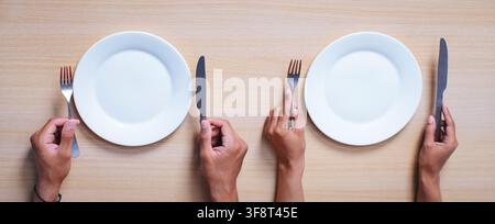 Unrecognizable Hands Holding Fork And Knife With Empty Plates On Wooden Table, Weight Loss Concept Stock Photo