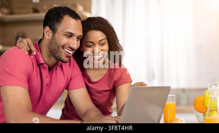 Happy Couple Using Laptop Together In Kitchen, Smiling And Enjoying Time Together Stock Photo