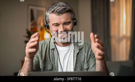 Smiling Mature Caucasian Man Using Laptop With Headset In Home Office, Business Concept Stock Photo