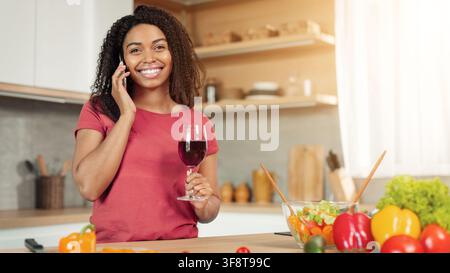 Happy African American Woman Talking On Phone And Holding Glass Of Red Wine In Kitchen Stock Photo
