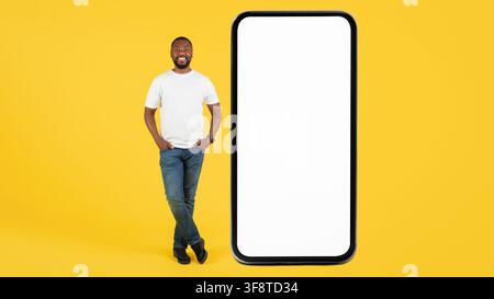 Smiling Black Man Standing Next To Smartphone With Blank Screen, Yellow Background, Advertising Concept Stock Photo