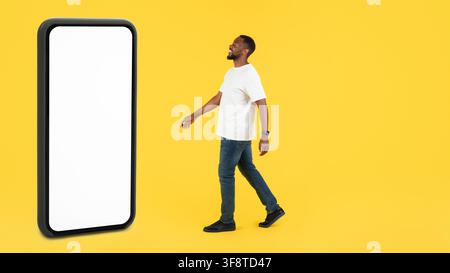 Happy Black Man Walking Towards Smartphone With Blank Screen, Yellow Background, Technology Concept Stock Photo