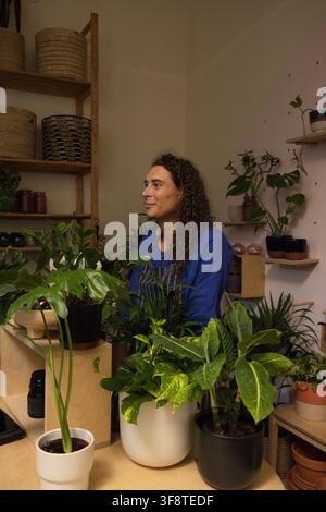 Non-binary worker in striped apron standing behind houseplants in plant shop arranging pots Stock Photo