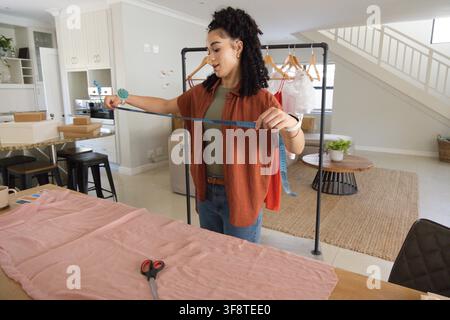 Female measuring light pink fabric at large table in open-plan living area using blue tape. Garment, sewing, studio, textile, workspace, home, fashion Stock Photo