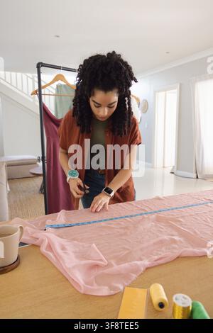 Woman measuring, marking pale-pink chiffon on table at home, in rust top, using blue measuring tape. Sewing, fabric, dressmaking, tailoring, studio, d Stock Photo