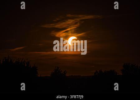 Partial solar eclipse seen through glowing orange clouds above a dark rural landscape in Lithuania, creating a dramatic atmospheric sky scene. Stock Photo