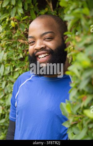 African American man smiling, standing by green hedge, wearing blue athletic shirt and black sleeve Stock Photo