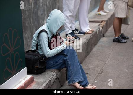 Woman using smartphone while sitting on sidewalk, side profile. Stock Photo