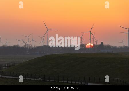 Glowing orange sun setting behind wind turbine farm and church spire with sheep grazing on green dike in Groningen Netherlands Stock Photo