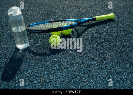 Tennis racket, yellow balls and water bottle in sunlight on outdoor hard court with copy space, sports training and hydration concept. Stock Photo