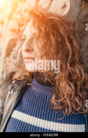 Woman curly hair portrait leaning against stone wall in warm golden sunset light creates a serene and thoughtful mood Stock Photo