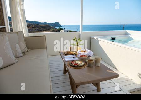 Enjoy a lavish brunch on a serene terrace in Milos, Greece. Overlooking sparkling blue waters and rugged cliffs, this scene captures the essence of re Stock Photo